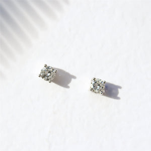 A product photo of a pair of 9 karat white gold lab diamond stud earrings sitting in the sun on a white textured background. The brilliant, colourless half pointer round diamonds measure approximately 4mm across, and are held in place by a simple 4-claw setting.