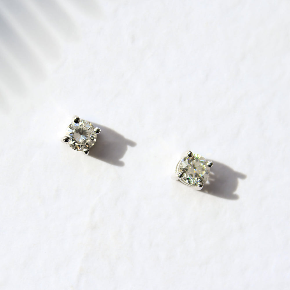 A product photo of a pair of 9 karat white gold lab diamond stud earrings sitting in the sun on a white textured background. The brilliant, colourless half pointer round diamonds measure approximately 3.5mm across, and are held in place by a simple 4-claw setting.