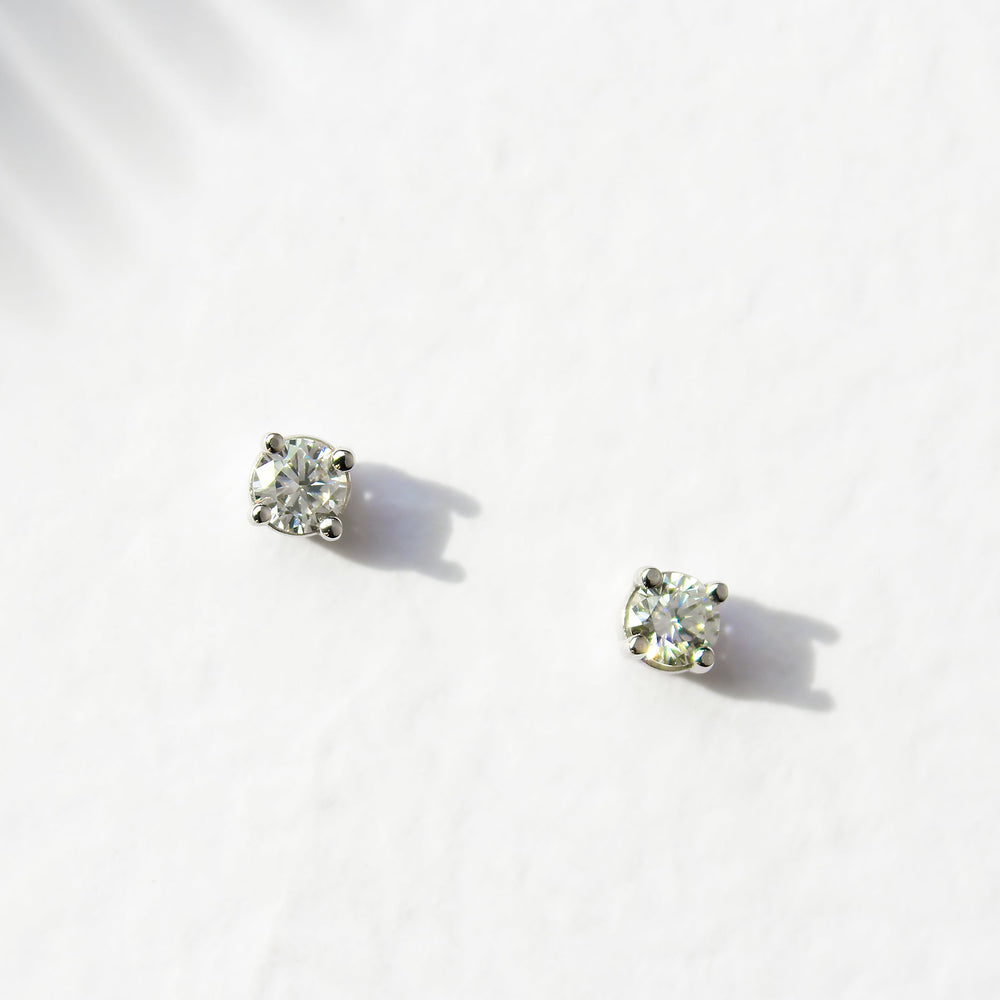 A product photo of a pair of 9 karat white gold lab diamond stud earrings sitting in the sun on a white textured background. The brilliant, colourless half pointer round diamonds measure approximately 3mm across, and are held in place by a simple 4-claw setting.