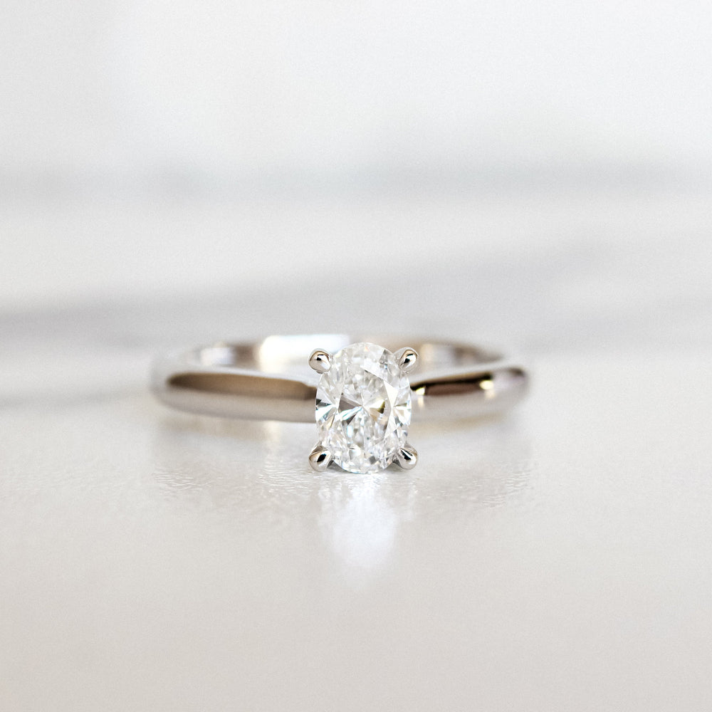 A product photo of a 14 karat white gold lab diamond solitaire engagement ring sitting in the sun on a white textured background. The brilliant, colourless half pointer oval-cut diamond measures 4.5mm across, and is held in place by 4 delicate claws in a high-profile trellis setting.