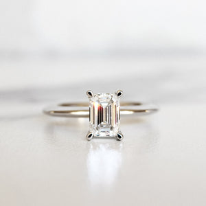 A product photo of a 14 karat white gold lab diamond engagement ring sitting in the sun on a white textured background. The brilliant, colourless 1ct emerald-cut diamond measures about 6.7mm lengthwise, and is held in place by 4 delicate golden claws in a prong setting.