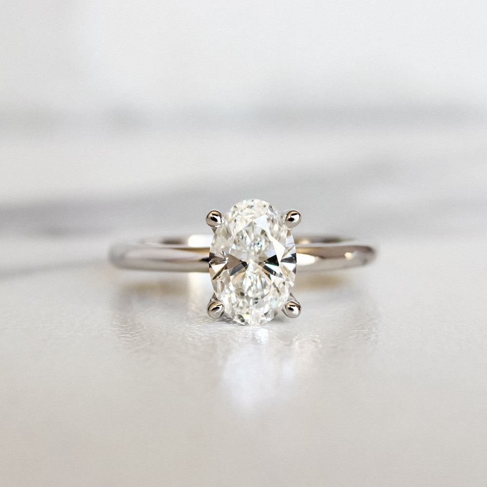 A product photo of a 14 karat white gold lab diamond solitaire engagement ring sitting in the sun on a white textured background. The brilliant, colourless 1 carat oval-cut diamond measures 8.4mm lengthwise, and is held in place by 4 delicate claws in a prong setting.