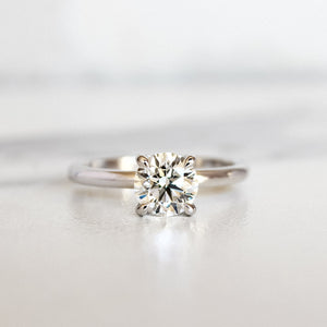 A product photo of a 14 karat white gold lab diamond solitaire engagement ring sitting in the sun on a white textured background. The brilliant, colourless 1 carat round-cut diamond measures 6.3mm lengthwise, and is held in place by 4 delicate claws in a prong setting.