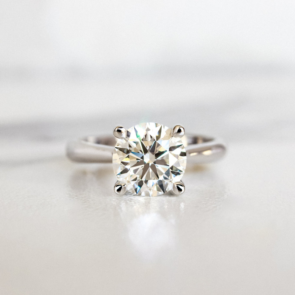 A product photo of a 14 karat white gold lab diamond solitaire engagement ring sitting in the sun on a white textured background. The brilliant, colourless 2 carat round-cut diamond measures over 8mm across, and is held in place by 4 delicate claws in a high-profile cathedral setting in the middle of the thick, tapered band.