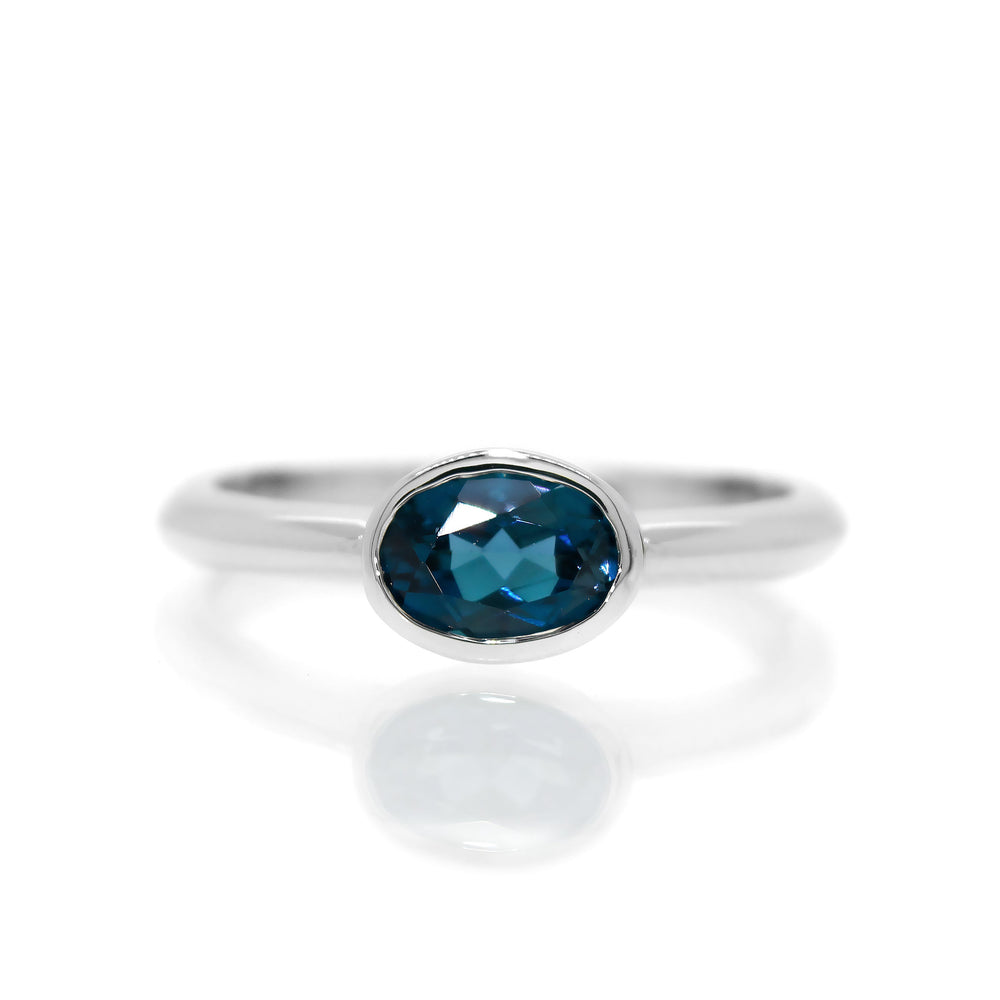A product photo of a silver ring with a bezel-set London Blue topaz centre stone sitting on a white background. The silver band is simple and smooth, connecting on either side of a horizontally-oriented oval-cut topaz stone surrounded by a solid frame of silver.