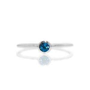 A product photo of a delicate silver stacking ring with a tiny, bezel-set London Blue Topaz in the centre sitting on a white background. The band is slim and thread-like, with the focus drawn to the petite 3mm glinting blue centre stone. The deep blue gemstone colour be a good sapphire alternative.