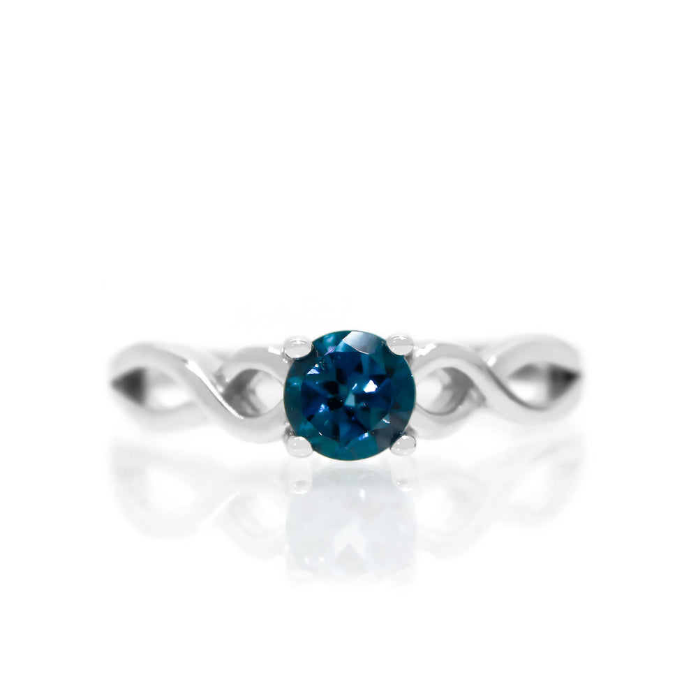 A product photo of an ornate silver ring with a london blue topaz centre stone sitting on a white background. The silver band splits halfway along its length, becoming twisting and serpentine in appearance before meeting on either side of the dazzling blue 5mm stone, which is held in place by 4 silver claws.