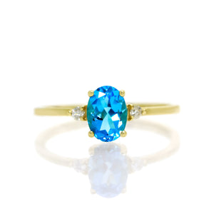 A product photo of a yellow gold blue topaz ring sitting against a white background. The yellow gold band is plain and smooth, and the centre oval-cut bright blue topaz stone is framed by a single white diamond on either side.