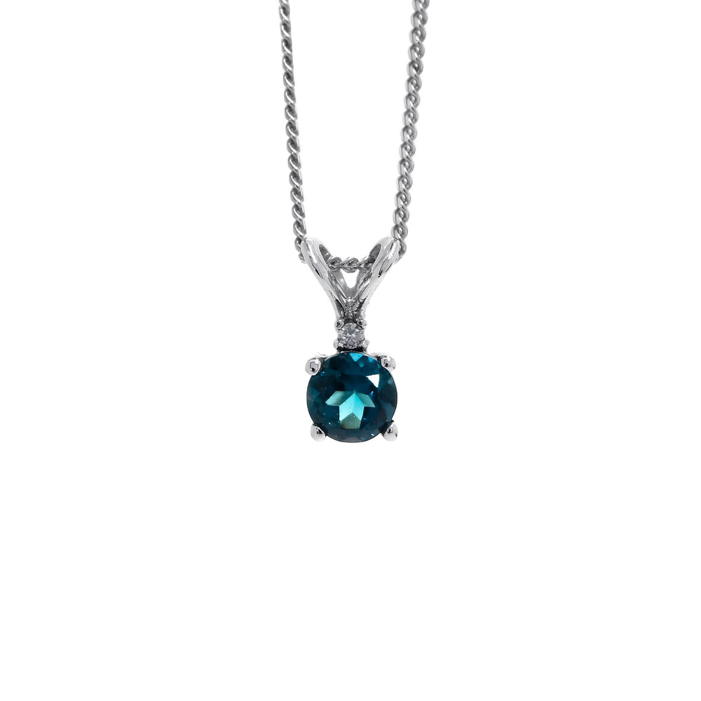 A product photo of a 4.5mm round London Blue Topaz and diamond pendant in 9k white gold suspended against a white background. The delicate topaz gemstone is a stunningly deep ocean blue colour, almost sapphire in appearance. A single white diamond sits atop it, before meeting the split golden bail.