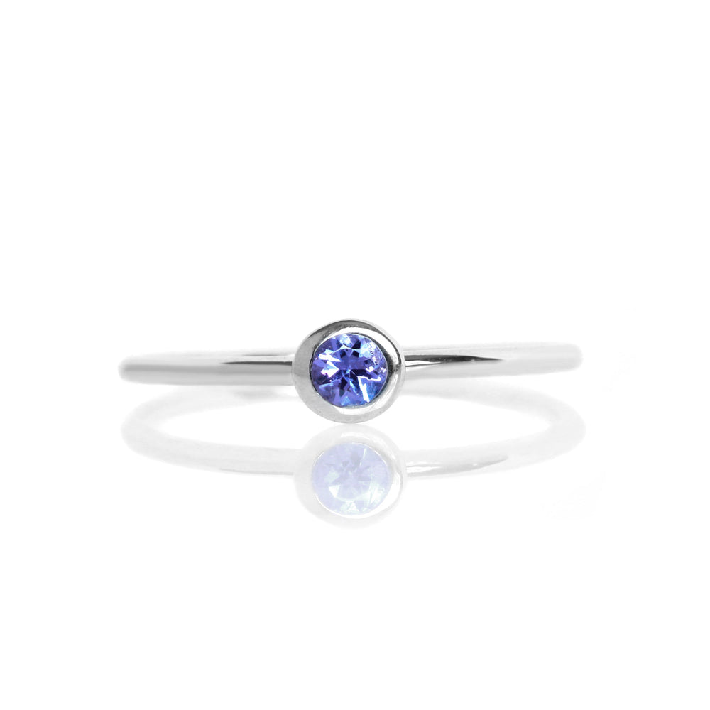 A product photo of a delicate silver stacking ring with a tiny, bezel-set tanzanite in the centre sitting on a white background. The band is slim and thread-like, with the focus drawn to the petite 3mm glinting blue centre stone. The deep blue gemstone colour would be a good sapphire alternative.
