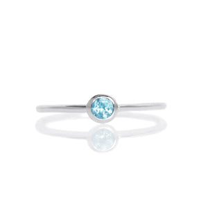 A product photo of a delicate silver stacking ring with a tiny, bezel-set blue topaz in the centre sitting on a white background. The band is slim and thread-like, with the focus drawn to the petite 3mm glinting blue topaz centre stone.