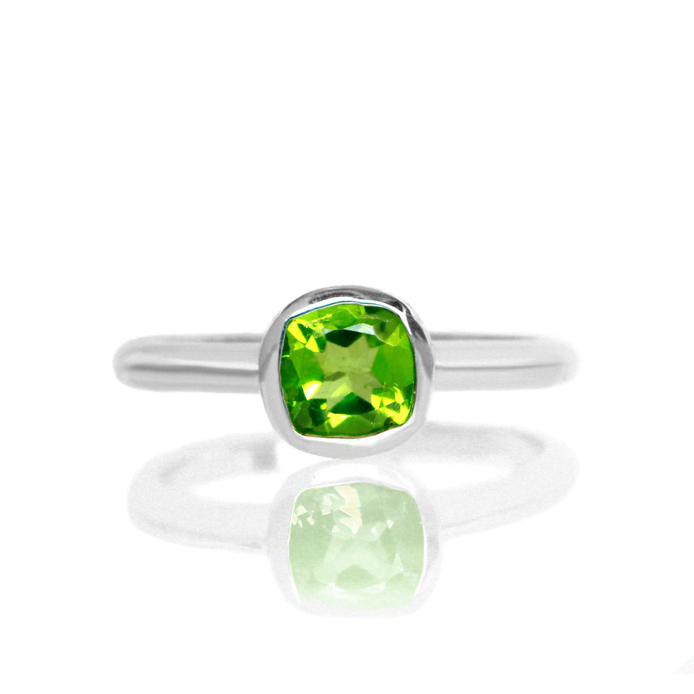 A product photo of a silver ring with a bezel-set peridot centre stone sitting on a white background. The silver band is simple and smooth, connecting on either side of a squared-cushion peridot stone surrounded by a solid frame of silver.