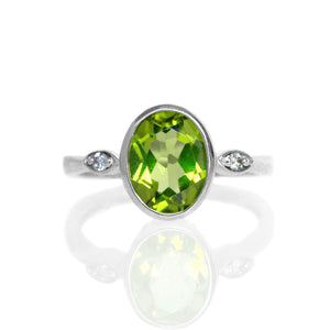 
            
                Load image into Gallery viewer, A product photo of a silver ring with a large bezel-set peridot centre stone sitting on a white background. The silver band is simple and smooth, connecting on either side of a vertically-oriented oval-cut peridot stone surrounded by a solid frame of silver. Before the band reaches the centre stone, it splits into two small horizontal silver oval frames on each side, each housing a small diamond.
            
        