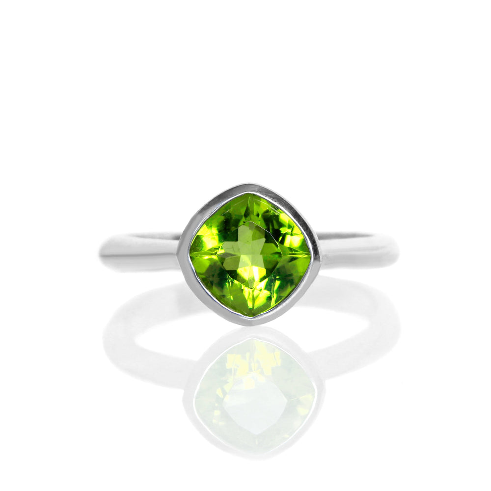 
            
                Load image into Gallery viewer, A product photo of a silver ring with a bezel-set peridot centre stone sitting on a white background. The silver band is simple and smooth, connecting on either side of a diagonally-oriented squared-cushion peridot stone surrounded by a solid frame of silver.
            
        