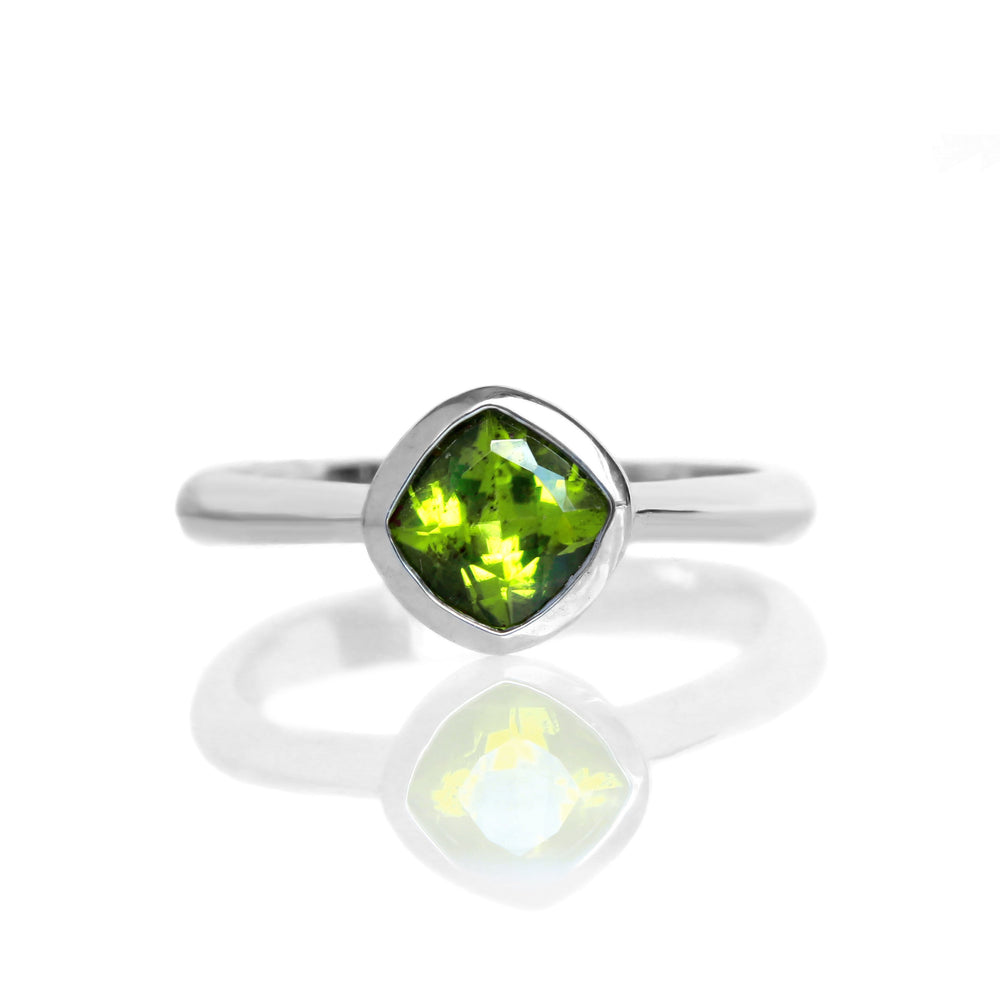 A product photo of a silver ring with a bezel-set peridot centre stone sitting on a white background. The silver band is simple and smooth, connecting on either side of a diagonally-oriented squared-cushion peridot stone surrounded by a solid frame of silver.