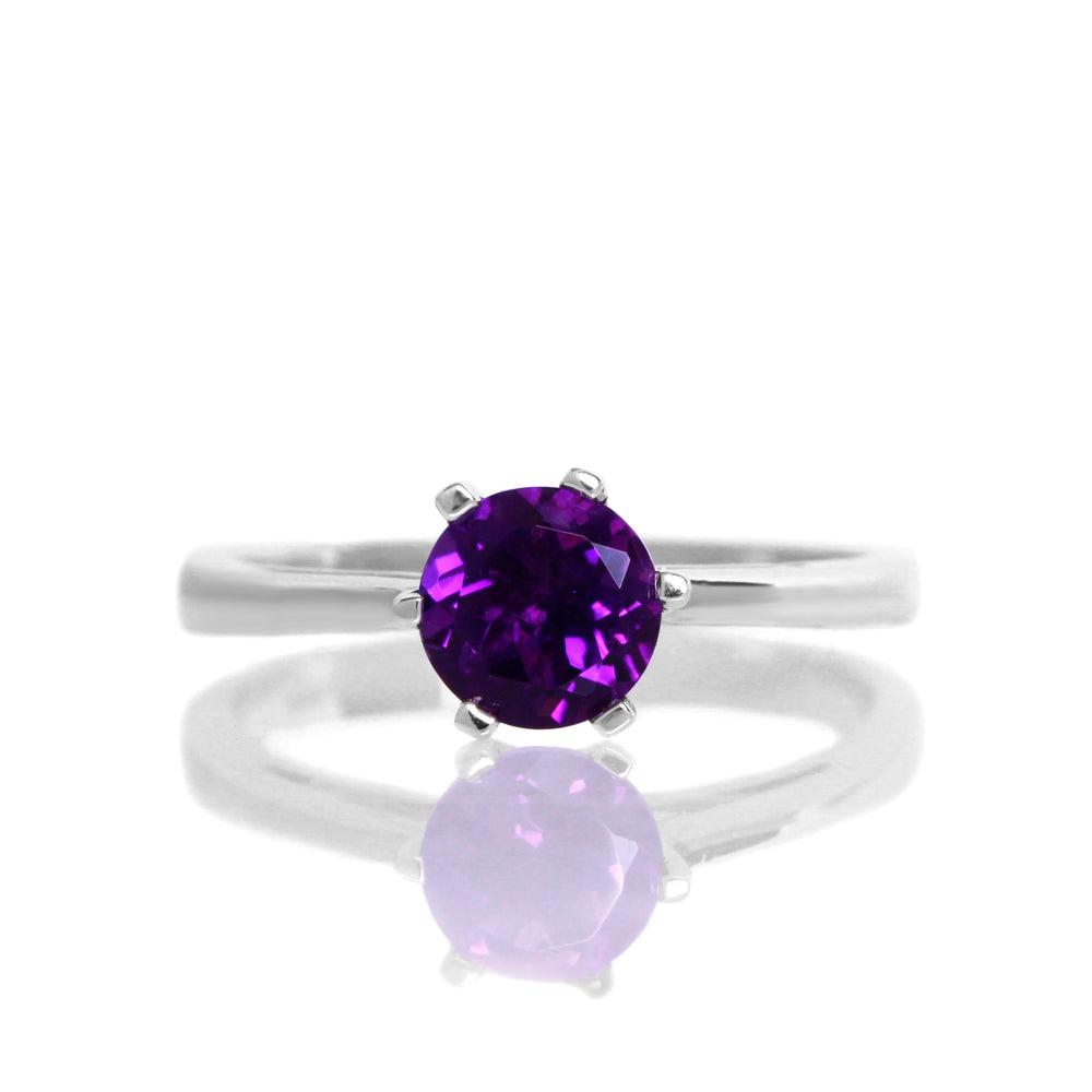 A product photo of a silver solitaire ring with an amethyst centre stone sitting on a white background. The silver band is simple and smooth, and the amethyst stone is held in place by 6 delicate silver claws.