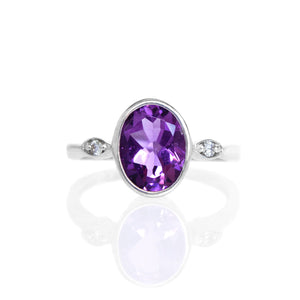 
            
                Load image into Gallery viewer, A product photo of a silver ring with a large bezel-set amethyst centre stone sitting on a white background. The silver band is simple and smooth, connecting on either side of a vertically-oriented oval-cut amethyst stone surrounded by a solid frame of silver. Before the band reaches the centre stone, it splits into two small horizontal silver oval frames on each side, each housing a small diamond.
            
        
