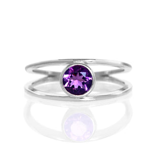 A product photo of a split-band silver ring with a bezel-set amethyst centre stone sitting on a white background. The silver band splits at the base of the ring, separating to meet at the top and bottom of the deep purple round-cut centre stone, framed in a thick layer of silver, holding it in place.