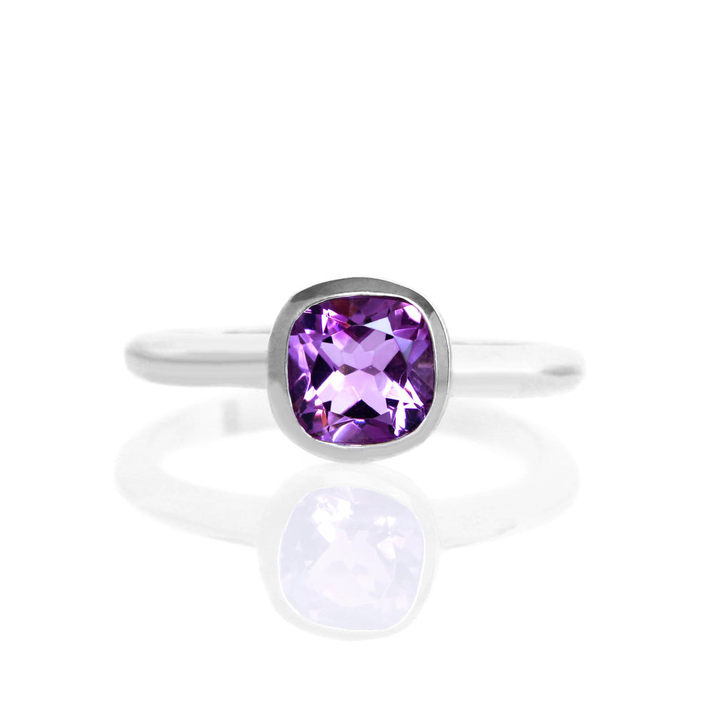 A product photo of a silver ring with a bezel-set amethyst centre stone sitting on a white background. The silver band is simple and smooth, connecting on either side of the squared-cushion purple amethyst stone surrounded by a solid frame of silver.