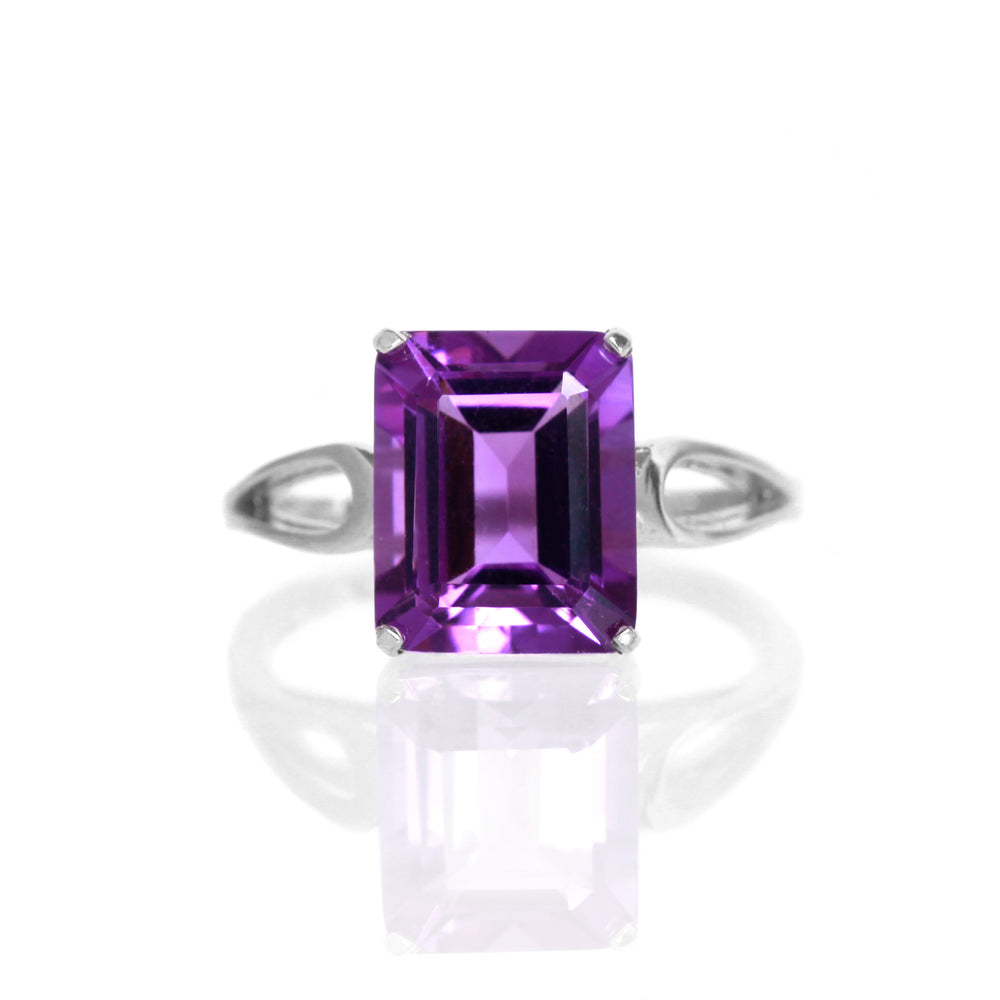 A product photo of an amethyst ring in silver sitting on a white background. The ring consists of a large, vertically-oriented rectangular centre stone, held in place by delicate silver loops in the band on either side. The jewel is a deep vibrant purple.