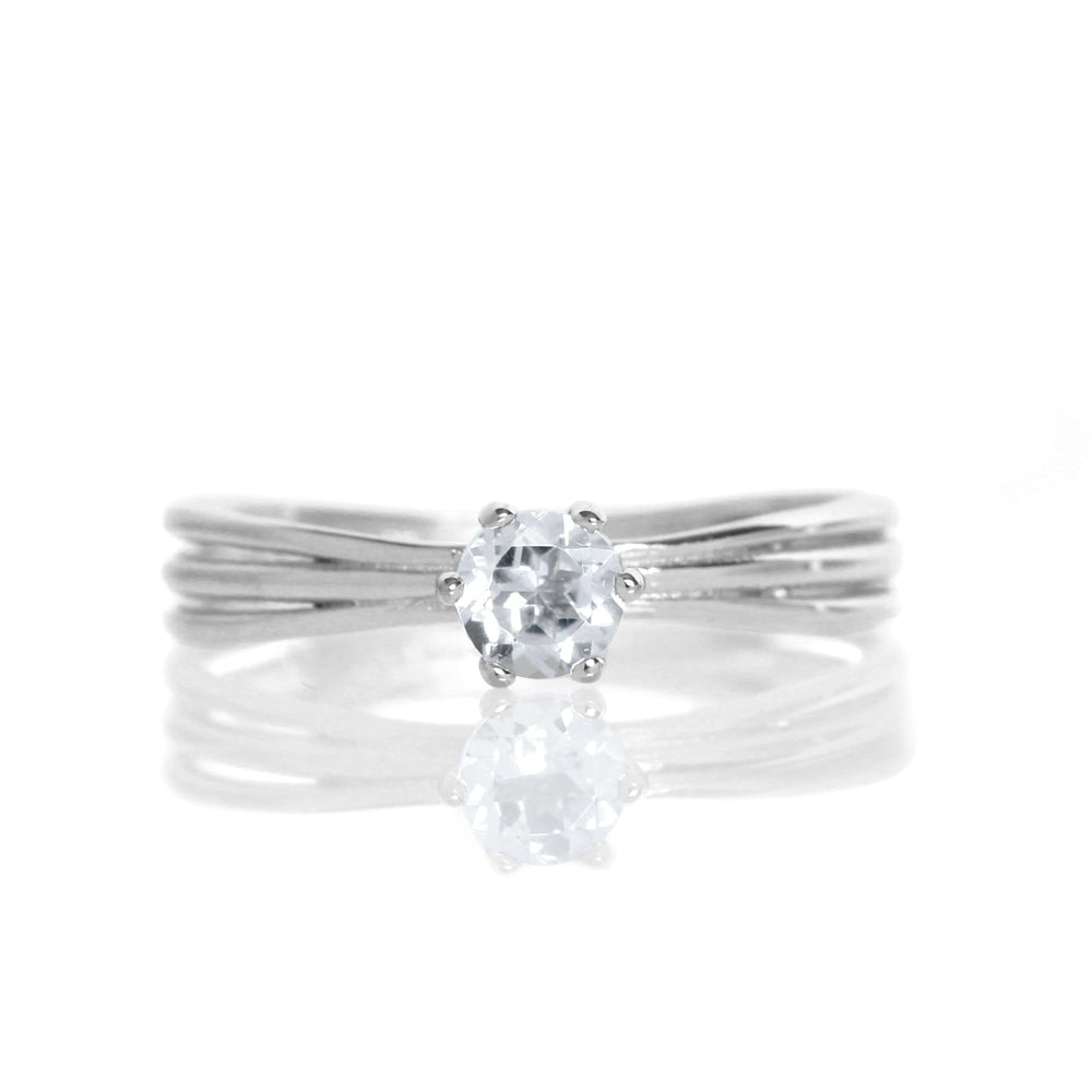 A product photo of a simple silver solitaire ring with a unique band detail and a 4mm round silver topaz centre stone sitting on a white background. The band is styled to appear as 3 little silver bands, tilted slightly outwards and "overlapping" towards the back of the ring.