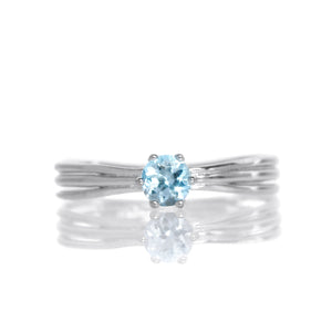A product photo of a simple silver solitaire ring with a unique band detail and a 4mm round aquamarine centre stone sitting on a white background. The band is styled to appear as 3 little silver bands, tilted slightly outwards and "overlapping" towards the back of the ring.