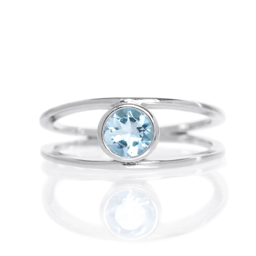 A product photo of a split-band silver ring with a bezel-set aquamarine centre stone sitting on a white background. The silver band splits at the base of the ring, separating to meet at the top and bottom of the pale blue round-cut centre stone, framed in a thick layer of silver, holding it in place.