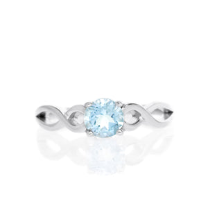 A product photo of an ornate silver ring with an aquamarine centre stone sitting on a white background. The silver band splits halfway along its length, becoming twisting and serpentine in appearance before meeting on either side of the dazzling blue 5mm stone, which is held in place by 4 silver claws.