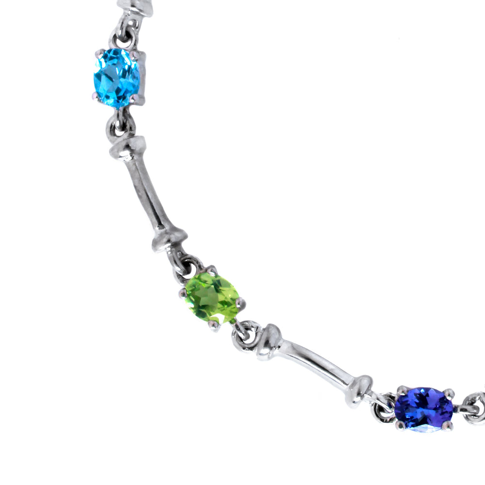 A product photo made up of a silver multi-gemstone bracelet featuring 4x5mm oval Blue Topaz, London Blue Topaz, Tanzanite and Peridot, all interspersed with solid silver bars.