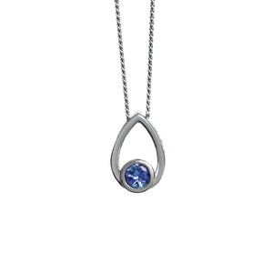 A product photo of a silver pendant with a single, tiny tanzanite stone encased in silver suspended against a white background. The bezel-set tanzanite sits at the bottom of a silver frame, shaped like a teardrop.