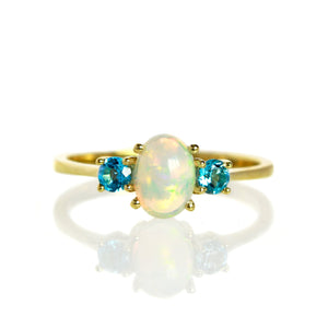 A product photo of a cabochon rainbow opal and blue topaz ring in 9 karat yellow gold on a white background. The cabochon cut of the opal allows the viewer to see every angle of the glittery fire within the translucent stone, while the blue topaz adds a burst of colour.
