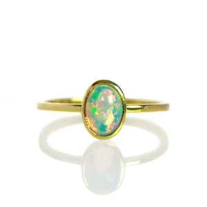 A product photo of a cabochon rainbow opal solitaire ring in 9 karat yellow gold on a white background. The cabochon cut of the opal allows the viewer to see every angle of the glittery fire within the translucent stone, which is held in place by a delicate bezel setting.