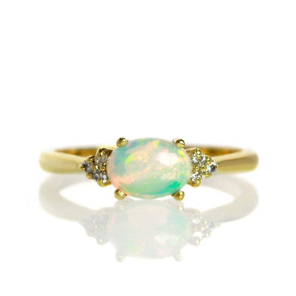 A product photo of a yellow gold cabochon rainbow opal and diamond ring sitting against a white background. The yellow gold band is plain and smooth, and the horizontally-oriented oval cabochon cut stone is framed by a delicate trio of white diamonds on either side.
