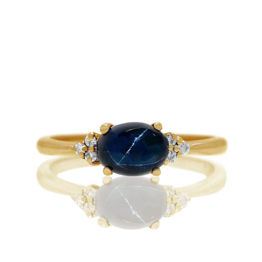 
            
                Load image into Gallery viewer, A product photo of a yellow gold cabochon star sapphire and diamond ring sitting against a white background. The yellow gold band is plain and smooth, and the horizontally-oriented oval cabochon cut stone is framed by a delicate trio of white diamonds on either side.
            
        