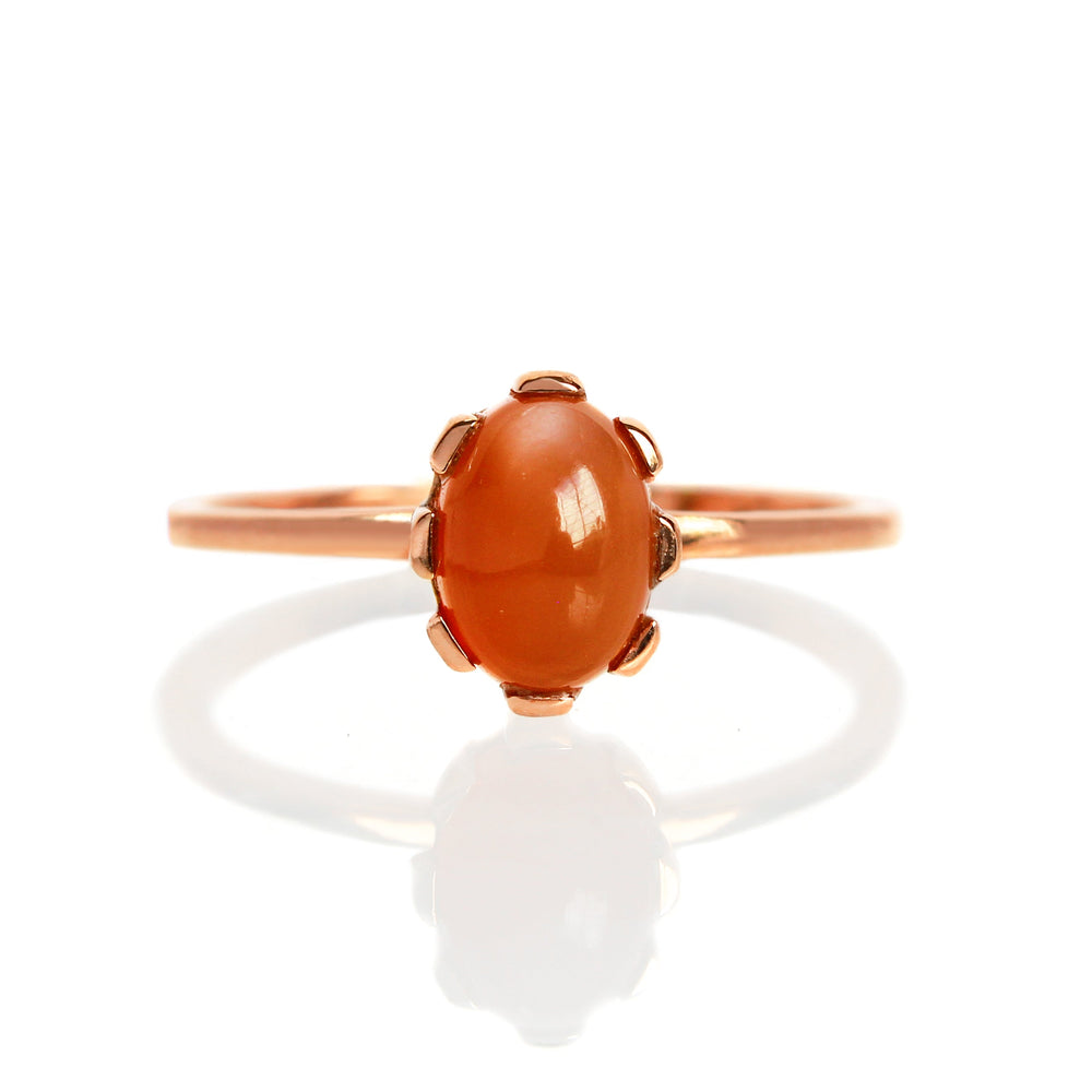 A product photo of a 7x5mm cabochon orange moonstone solitaire stacking ring in 9k rose gold. The ring's band is delicately thin, and flattened horizontally. The orange moonstone is held in place by a unique multi-claw setting.
