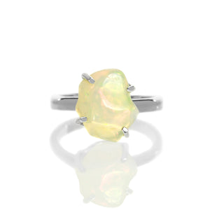 A product photo of a boho style statement ring on a white background. The ring is made up of a thick, flat sterling silver band, and a rough-cut rainbow opal gemstone. The stone is large and reflects a multitude of bright colours in irregular shapes.