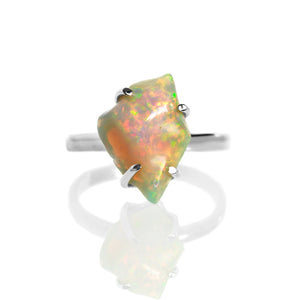A product photo of a boho style statement ring on a white background. The ring is made up of a thick, flat sterling silver band, and a rough-cut rainbow opal gemstone. The stone is large and reflects a multitude of bright colours in irregular shapes.