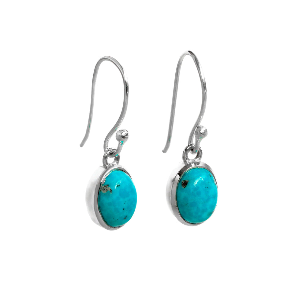 A product photo of a pair of sterling silver turquoise drop earrings suspended against a white background. The drop earrings feature shepherd hooks, and the 9x7mm oval-shaped cabochon turquoise gemstones are held in place by silver bezel settings. The stones are sparsley decorated by interesting dark natural inclusions.