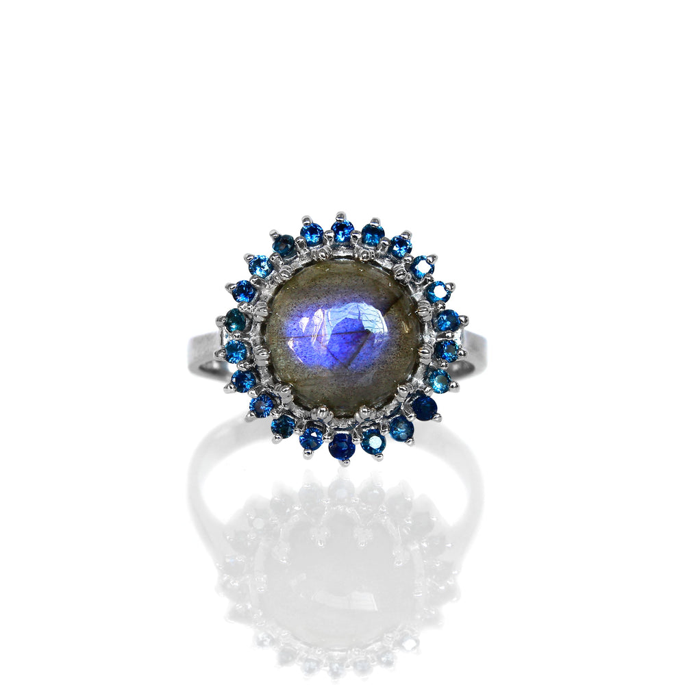 A product photo of a white gold statement ring sitting on a white background. The ring features a grey 9mm cabochon Labradorite gemstone with bright blue opalescent fire, surrounded by a halo of 22 blue sapphire jewels.