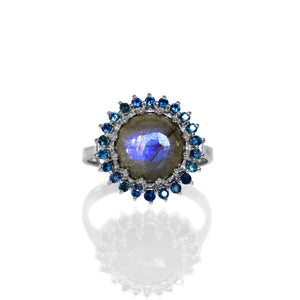 
            
                Load image into Gallery viewer, A product photo of a white gold statement ring sitting on a white background. The ring features a grey 9mm cabochon Labradorite gemstone with bright blue opalescent fire, surrounded by a halo of 22 blue sapphire jewels.
            
        