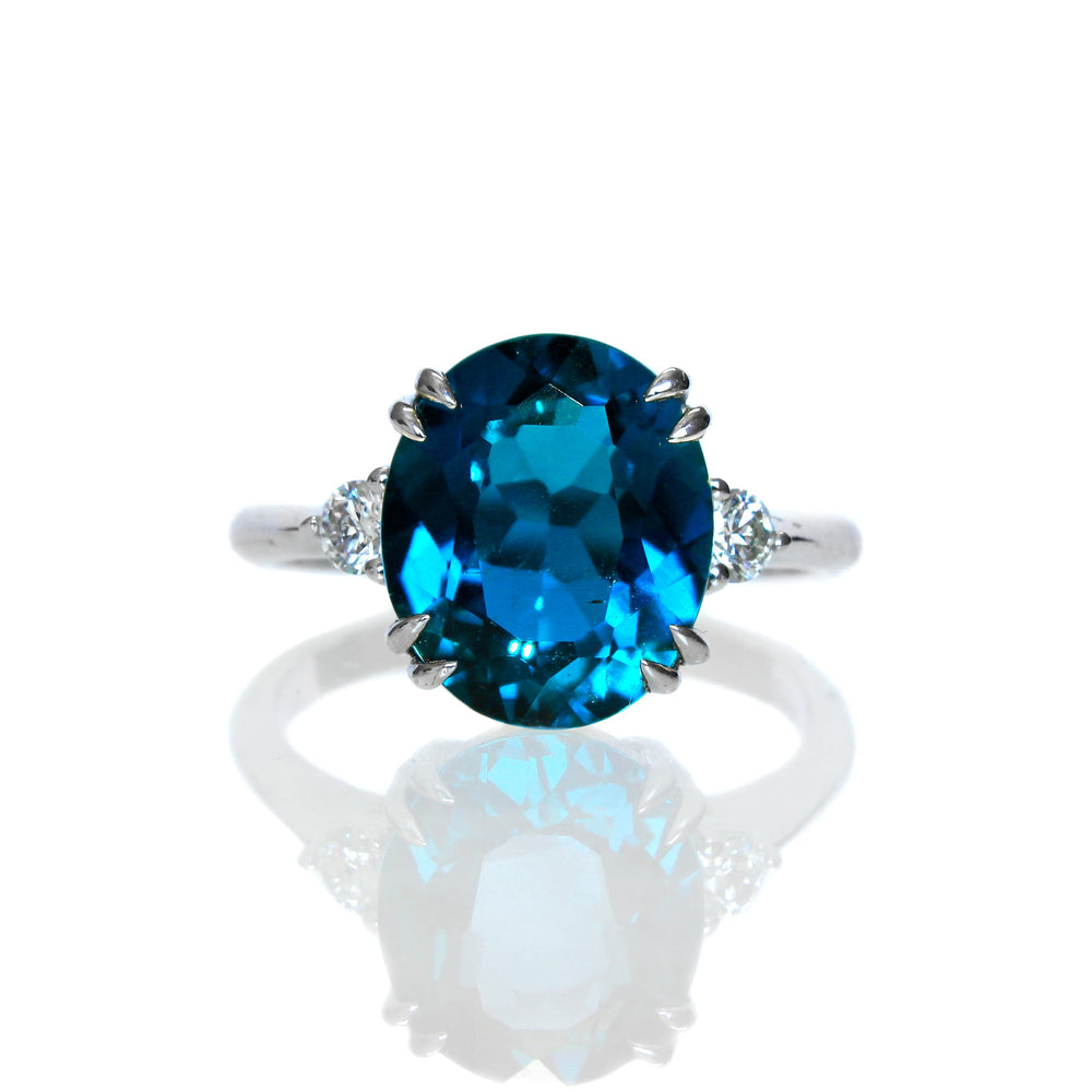 A product photo featuring a silver diamond and London Blue Topaz ring sitting on a white background. The London Blue Topaz is impressively large at a whole 3 carats, and is shaped in a faceted oval cut. Two round white diamonds, each weighing 0.10ct, sit on either side. The silver band is smooth and round.