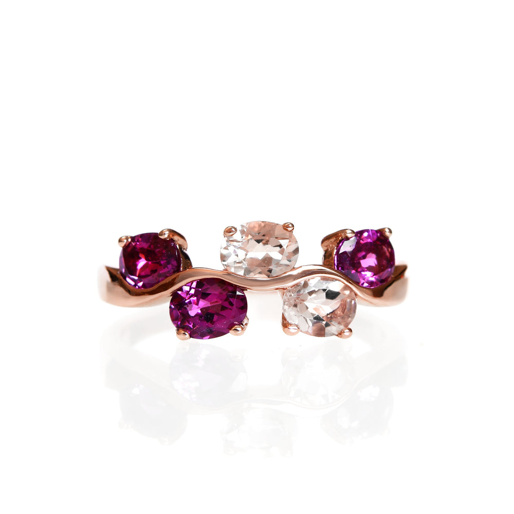 A product photo of a unique rose gold ring decorated with 5 separate grape garnet and morganite jewels. The rose gold band twists and curves elaborately, while the vibrant purple and pale pink gemstones decorate its length at regular intervals, appearing like grapes on a silver vine.