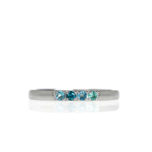 A product photo of a minimalistic 9 karat white gold stacking ring on a white background. The ring's band is smooth with a slight bevelled-edge, and has 4 2mm round blue gemstones embedded along its length - blue topaz, blue tourmaline, blue topaz and mint blue tourmaline.
