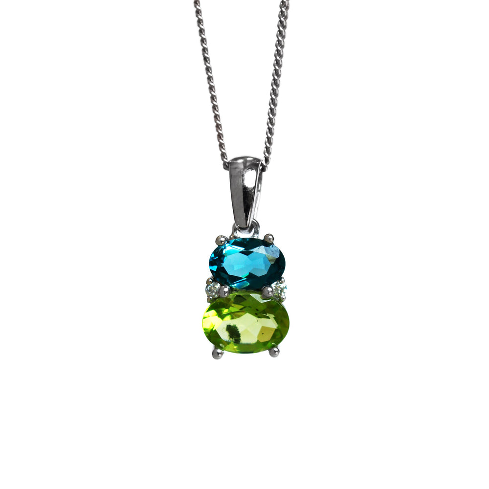 A product photo of a london blue topaz, peridot and diamond necklace in 9k white gold sitting on a white background. The pendant is made up of two horizontal oval-shaped bstones, the bottom one being a 7x5mm peridot and the top being a 6x4mm london blue topaz. The spaces between the two stones are filled with one small diamond on either side.