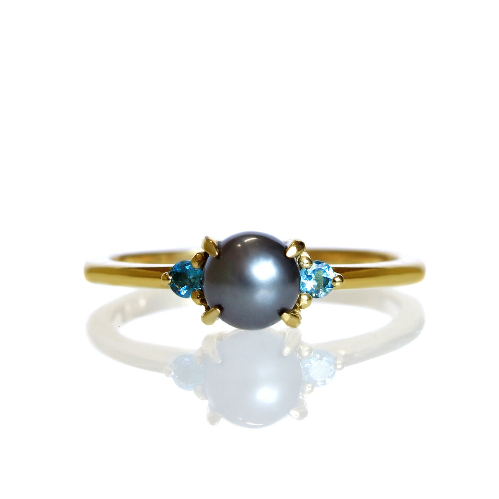A product photo of a solid 9 karat yellow gold pearl engagement ring sitting on a white background. The pearl is a 5mm perfectly spherical steel grey pearl, hugged on either side by a delicate aquamarine jewel.