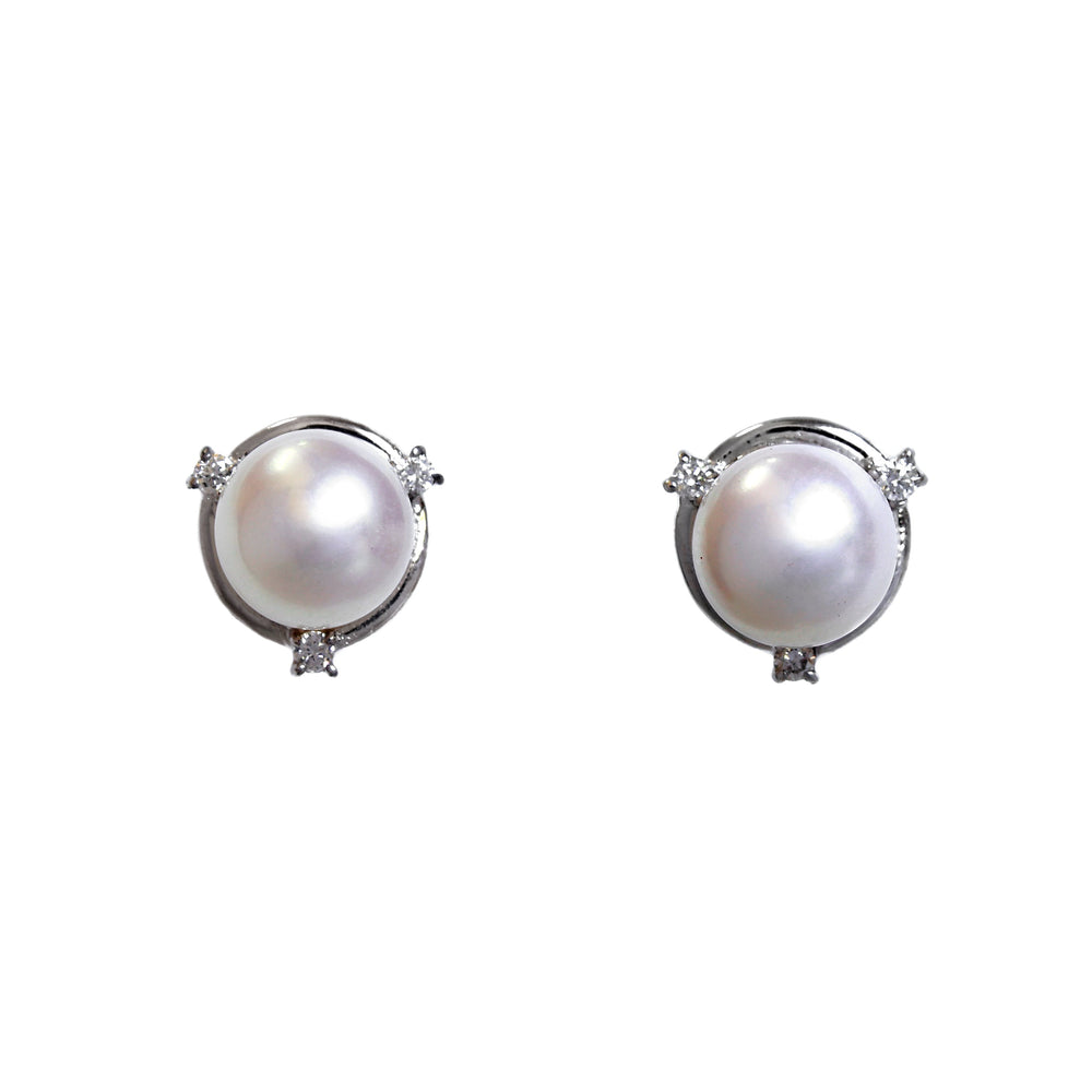 A product photo of a pair of white pearl and diamond stud earrings on a white background. The pearls are 7mm wide and perfectly round, and are set within swirling frames of solid white gold, featuring three tiny diamonds on each earring.