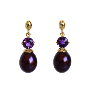 A product photo of a pair of solid yellow gold gemstone and pearl earrings with a unique design. The main components are two deep purple round-cut amethyst jewels, each one set above a single, dark peacock drop pearl, beautifully matching in colour.