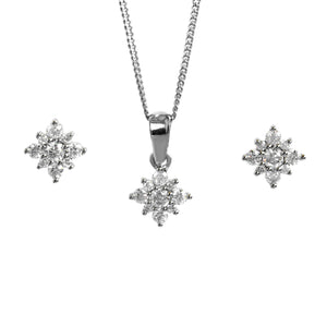 A product photo of a women's jewellery gift set in 9ct white gold and sparkling moissanite suspended against a white background. The white gold moissanite earrrings, each made up of glittering stones arranged in a floral or starlike formation, sit on either side of a pendant of the same design suspended by a silver chain.